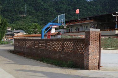 Çin 'in kırsal kesimindeki okulun tuğla duvarı üzerine kırmızı tuğlalar, beyaz duvarlar ve mavi süslemeler ve yanında da açık hava basketbol potası ile inşa edilmiştir. Arkaplanda yeşil dağlar ve ağaçlar yer alıyor.