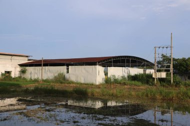 Demir çatılı beyaz beton bir bina, yağmurdan ve güneşten korunmak için plastik çarşaflarla kaplı pirinç tarlalarının ortasında yapım aşamasında..
