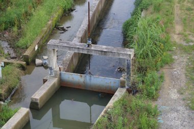 İçinden su akan açık ve biraz kirli beton bir kanalın manzarası. Merkezde, balıkların çitin altından geçmesini sağlayacak küçük bir metal kapı var..