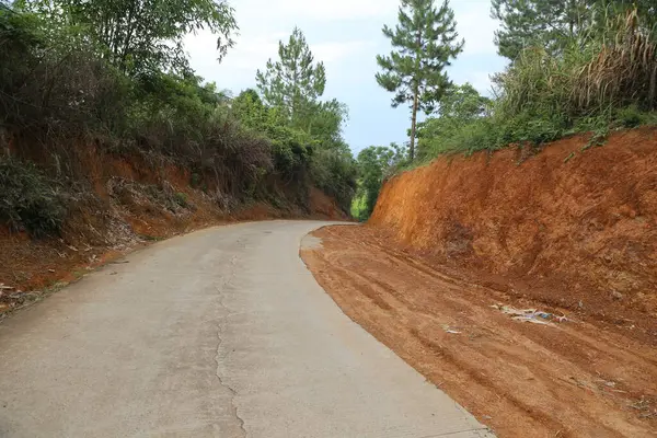Asfalt yolun kenarında, her iki tarafta da kırmızı toprak ve toprak var. Yamaçta farklı yüksekliklerde büyüyen ağaçlar var. Yolun bir ucunda da beton var.