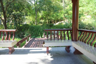 Yeşillik ve ağaçlarla çevrili, iki beyaz taş sıralı, geniş ahşap bir teras. Perspektif, bir bankın sol tarafından, yukarıdaki yüksek bir platformda benzer bir kürsüye bakıyor..