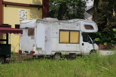Eski, beyaz ve yıpranmış bir karavan çimenli bahçedeki bir apartmanın önünde park edilmiş. Römork toz ve pasla kaplı, pencerelerinde sarı perdeler asılı..