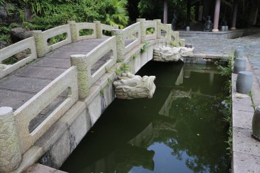 Ejderha başlı parmaklıklı taş bir köprü, önündeki yeşil bitkiler ve ağaçlarla çevrili küçük havuza götürür. Su yüzeyi bariyerlerin her iki tarafına da güneş ışığı yansıtıyor..
