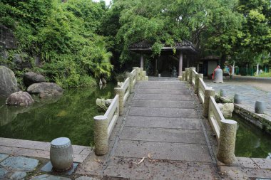 Yeşil ağaçlar ve suyla çevrili taştan bir köprü, bir tarafında büyük bir gölet bulunan pavyona götürür. Kamera, yakın bir yerden omuz üstünden bir perspektif yakalıyor..