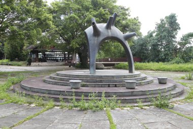 Yuvarlak bir platform, ona doğru giden adımlar, üzerinde iki el birbirine sarılıp üst üste oturan soyut bir heykel var. Arka planda yeşil ağaçlar, otlar, beton zeminler ve tuvaletteki binalar yer alıyor.