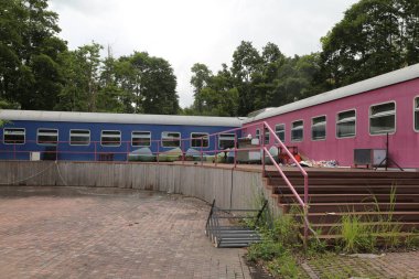 İki tren vagonu, yan yana, biri mavi ve diğeri pencereli, küçük bir açık alanda kaykay parkı olarak kurulmuş, park alanının içinde, ağaçlarla çevrili.