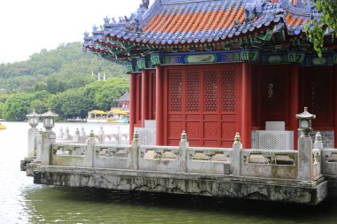 Suda kırmızı ve beyaz bir taş bina, Çin mimarisi tarzında koyu mavi kiremitleri ve kırmızı duvarları var. Önünde yeşil ağaçlarla çevrili klasik mimari tarzda antik bir pavyon var.