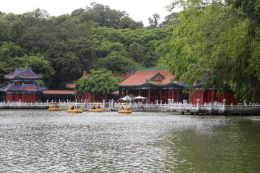 Çin mimarisi tarzında kırmızı ve beyaz bir bina, yeşil ağaçlarla çevrili gölün üzerinde duruyor. Önünde açık hava pavyonu ve suda yüzen küçük sarı kayıklar var..