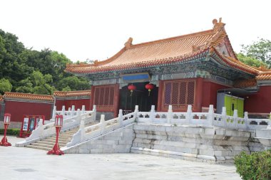 Sol tarafında yeşil ağaçlarla çevrili iki basamaklı kırmızı ve beyaz bir taş bina. Çatı mavi karolarla kaplı ve geleneksel desenlerle süslenmiş..