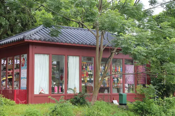Parkta kırmızı Kore tarzı bir dükkan. İki duvarın bir tarafında renkli ürünlerin olduğu büyük bir pencere. Yanında yaşlı bir ağaç var. Çatıda siyah fayanslar var. Yaz mevsimindeyiz ve etrafta yeşil çimenler var..