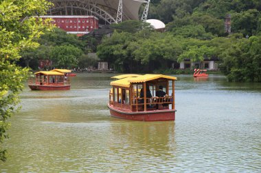 Sarı çatılı kırmızı ahşap bir Çin teknesi ve açık hava parkındaki bir gölde yüzen insanlar. Sahneyi çevreleyen ağaçlar ve arka planda büyük bir stadyum binası bulunuyor.