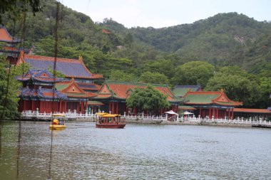 Sahilde bir grup kırmızı ve yeşil bina, Çin tarzı mimari, beyaz duvarlar ve mavi fayanslar. Önünde göl yüzeyinde yüzen antik bir tekne var..