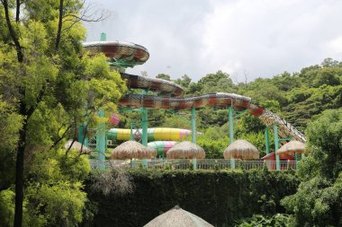 Yeşil ağaçlarla çevrili bir tepenin tepesinde ve sazdan çatı kulübeleri olan piton şeklinde bir su parkı. Su kaydıraklarının aşağıdaki çeşitli havuz alanlarına giden renkli boruları var..