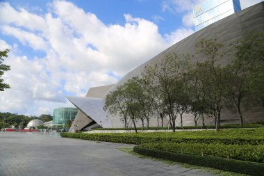 A large building with a lot of trees in front of it. The building is very tall. There are a few people walking around the area