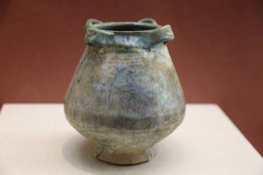 in the museum, an ancient chinese blue-green ceramic wine jar is placed on top of a white cloth with four feet at its base and three ear-shaped handles around it.