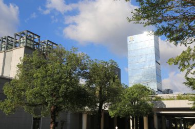 a tall building with glass curtain walls and trees in front of it, the top floor is decorated as an art installation for figures to stand on. the sky above has white clouds and blue tones.