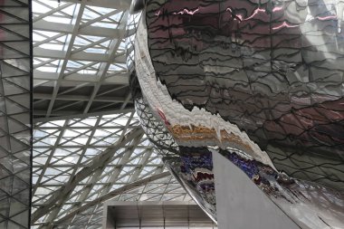 exterior close-up of a massive, intricate, scaly silver and rose gold metal geometric structure with angular steel structures in the background.
