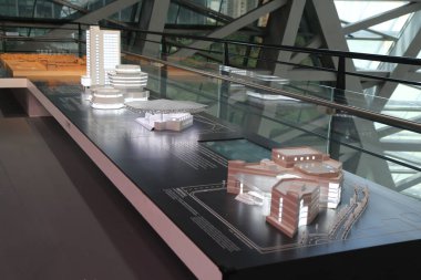 a black glass table displays architectural models of buildings in the city, with some model parts placed on it. the background is an open space inside the exhibition hall, featuring large floor-to-ceiling windows and gray metal railings