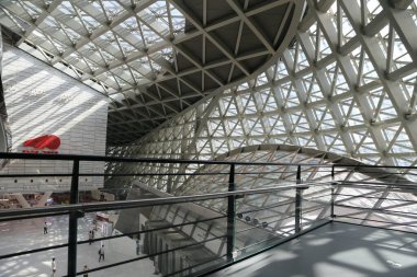 the indoor space of the glass and steel architecture features an all-white metal structure with triangular grid elements, forming complex geometric shapes that create visual effects through light refraction in sunlight