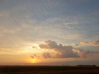 Yolun ortasındaki kırsal alan, ağaçlar, çalılar ve güzel bir günbatımı boyaları çok renkli gökyüzü ve dev bulutlar gökyüzünü tekeline alıyor.. 
