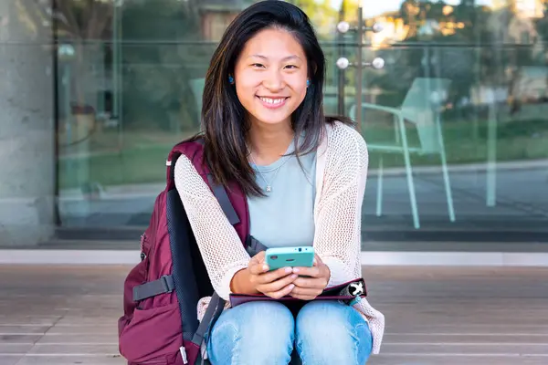 Beautiful Chinese teen student smiling using social media app on mobile phone outdoors. Girl sitting chatting on mobile phone