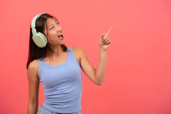 一位年轻美丽的亚洲女子的画像 她戴着耳机 一只手拿着手指 左上方挂着笑脸 站在演播室里的女孩广告和展示概念 — 图库照片