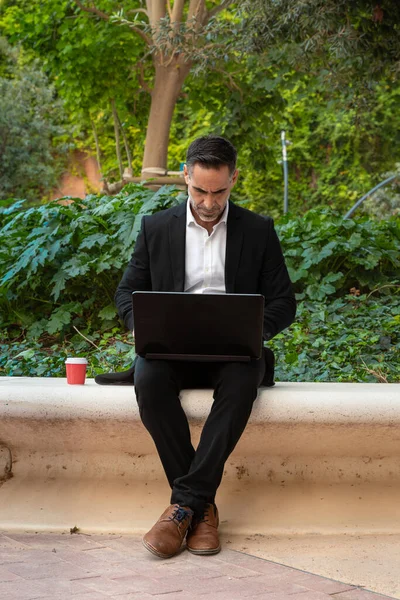 A middle-aged adult man in a suit has a business call while looking at his laptop.