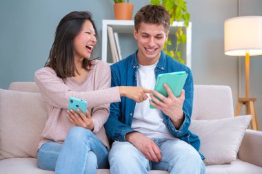  Genç, gülümseyen, çok kültürlü bir çift evdeki koltukta, bir cihazdaki kablosuz internet uygulamasını tarayan bir tablet ve cep telefonu kullanarak oturuyor. Teknoloji konsepti.