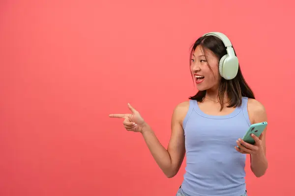 ヘッドフォンを着ている若いアジア人女性は 驚いた顔で写真の左側を指差します スタジオにスマートフォンを持って立っている少女 広告とプレゼンテーションの概念 — ストック写真