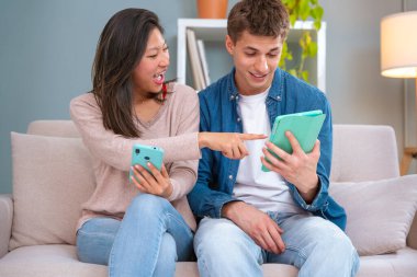  Genç, gülümseyen, çok kültürlü bir çift evdeki koltukta, bir cihazdaki kablosuz internet uygulamasını tarayan bir tablet ve cep telefonu kullanarak oturuyor. Teknoloji konsepti.