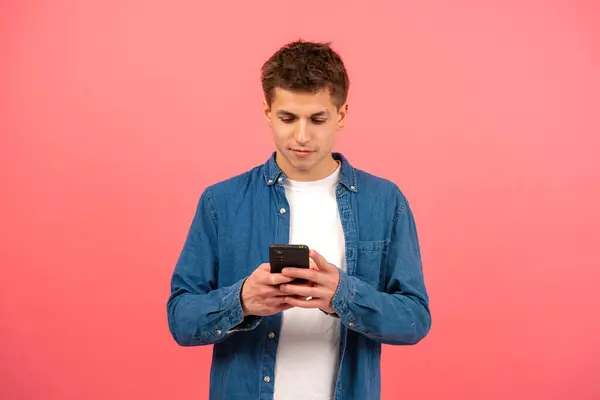 Portrait of happy man looking at mobile phone, smiling, texting over pink background. Concept of human emotions, facial expression. Copy space for ad