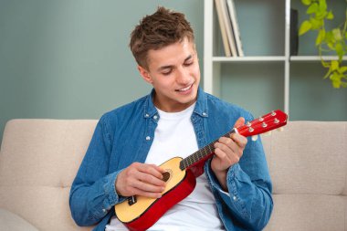 Genç adam evde ukulele çalıyor. Medya. Adam evde tek başına ukulele çalıyor. Genç adam kanepede otururken küçük bir gitar çalma pratiği yapıyor.