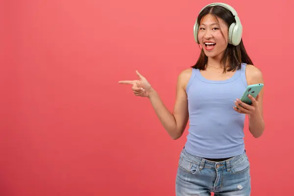 年轻的亚洲女人戴着耳机 手指指向照片的左边 脸上带着惊讶的表情 那个站在演播室里拿着电话的女孩 广告和展示的概念 — 图库照片