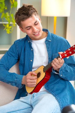 Genç adam evde ukulele çalıyor. Medya. Adam evde tek başına ukulele çalıyor. Genç adam kanepede otururken küçük bir gitar çalma pratiği yapıyor.