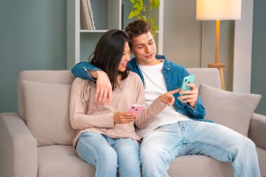  Genç, gülümseyen, çok kültürlü bir çift evdeki koltukta oturuyor cep telefonuyla bir cihazdaki kablosuz internet uygulamasını tarıyor. Teknoloji konsepti.