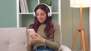 Kulaklıklı güzel bir kadın kolay ve güvenli elektronik ödemeler yapmak için kanepede mutlu bir şekilde internetten cep telefonuyla alışveriş yapıyor. İçeride.