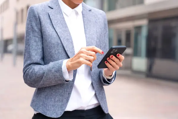 Satisfied with the results, the businesswoman walking on the street outside the office building, a mature boss holds a phone in her hands, writes messages and reads news online, using an app.