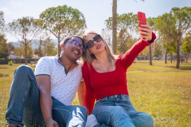 Güneş gözlüklü mutlu çift cep telefonuyla selfie çekiyor. Öğleden sonra parkta eğleniyorlar..