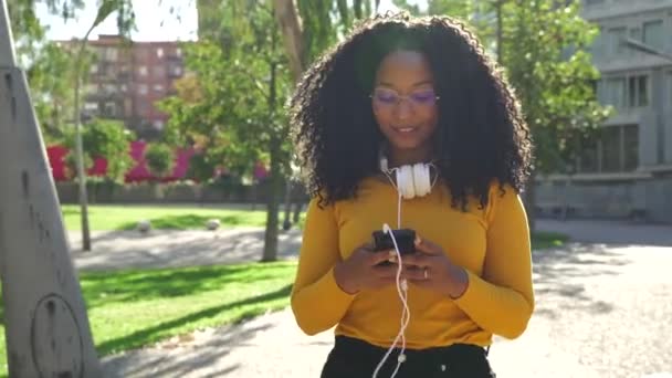 Piękna Czarna Kobieta Kręconymi Włosami Słuchawki Spacery Podczas Korzystania Smartfona — Wideo stockowe