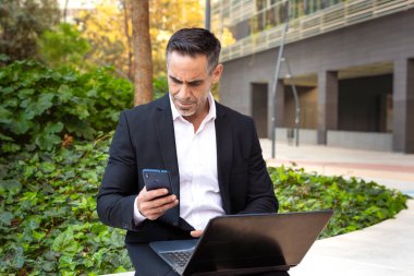 Takım elbiseli yetişkin bir adam elinde bir telefon tutuyor, mesajlar yazıyor ve internetten bir uygulama kullanarak haberleri okuyor.
