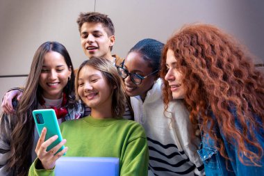 Beş çok ırklı arkadaş üniversite kampüsünde sosyal medyayı akıllı telefonlarıyla kullanırken eğleniyor. Üniversite yaşam konsepti.