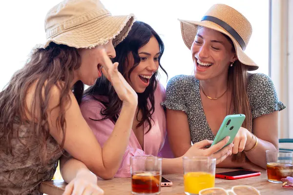 Bir grup gülen etnik kadın bir restoranda tatilin tadını çıkarıyor. Güzel ve neşeli Z Nesli kızları internette akıllı telefonlarıyla geziniyor.