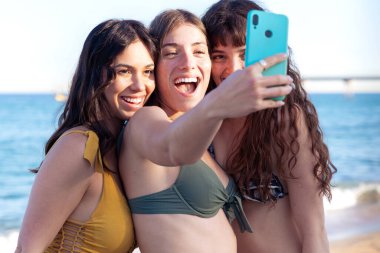 Mayo giymiş, gülümseyen, etnik çeşitliliğe sahip bir grup kadın tatilin tadını çıkarıyor. Z neslinin güzel ve neşeli kızları cep telefonlu bir fotoğraf için poz veriyor..