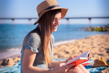 Sahilde oturan, şapkalı, arka planda deniz olan bir kadın kitap okuyor.