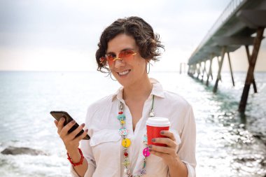 Sahilde elinde cep telefonu olan, güneş gözlüklü gülümseyen bir kadın portresi.