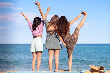 Sahilde mutlu bir şekilde el ele tutuşan neşeli mayo giymiş çok ırklı kadın arkadaşlar.