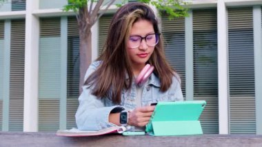 Kulaklıklı genç bir kız öğrenci, açık havada elinde dijital tablet ve kredi kartıyla online alışveriş yapıyor. Konsept: Çevrimiçi alışveriş.