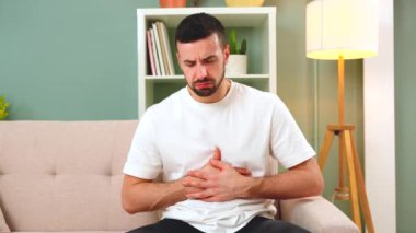 The man is sitting on a gray couch and holding his belly. Medicine and health concept, stomach problems. The man suffers from stomach ache, gastric problems. Abdominal pain, suffering and pain.