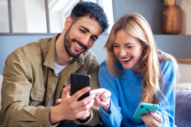 Şaşırmış bir kadın ve akıllı telefona bakan gülümseyen bir adam birlikte heyecanlı bir anı paylaşıyorlar. Her ikisi de ellerinde telefonlar, evde oturuyorlar, neşeli bir ifade ve harika adamlar.