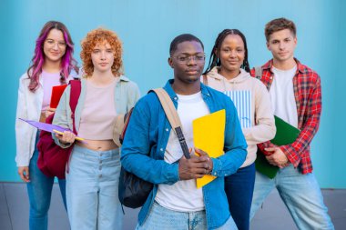 Üniversite kampüsündeki bir grup öfkeli öğrenci ellerinde not defterleriyle mavi arka planda kameraya bakıyor.